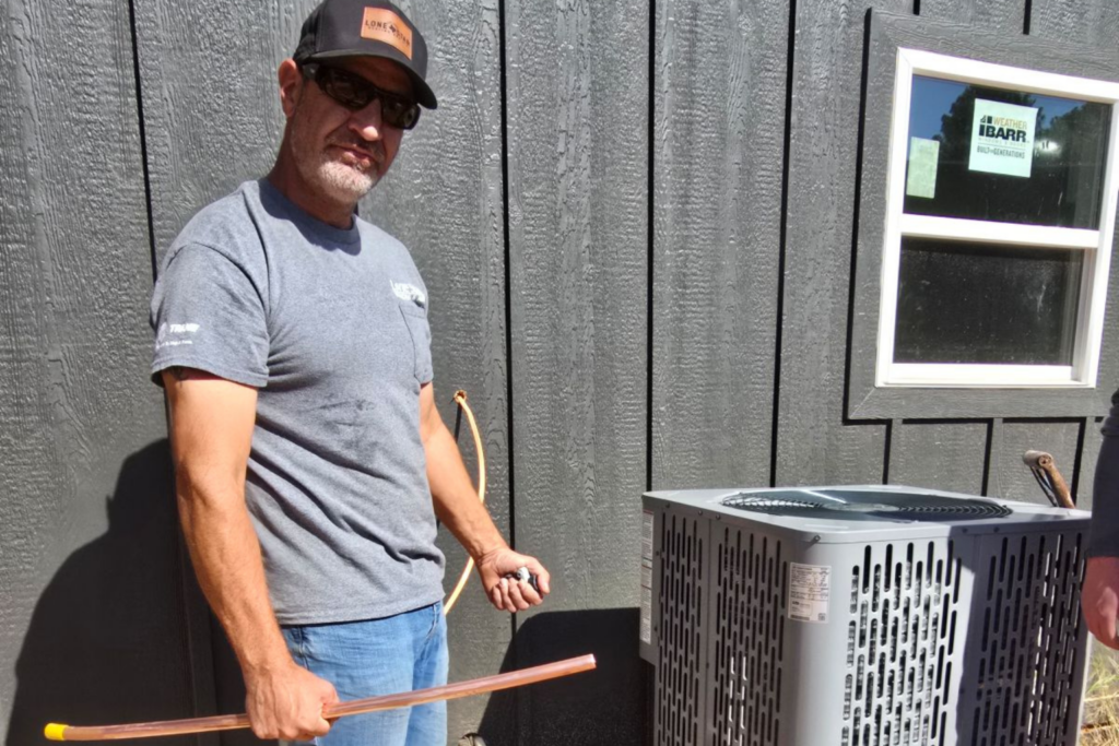 A technician wearing a grey t-shirt, sunglasses, and a "Lone Star" branded hat stands next to a newly installed grey AC condenser unit. He is holding a length of copper piping in front of a dark, wood-paneled exterior wall.