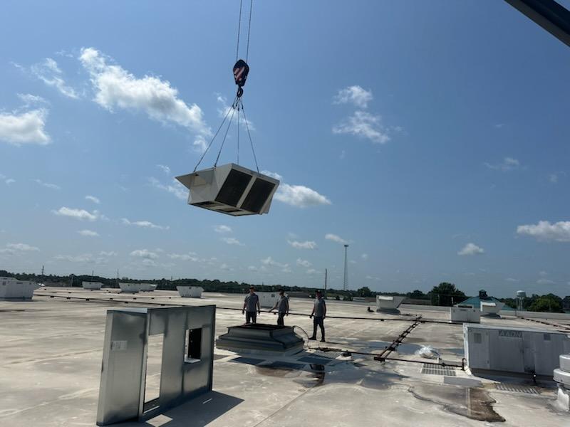 A crane hoists a large HVAC unit over a flat commercial roof where three workers stand ready to position it.
