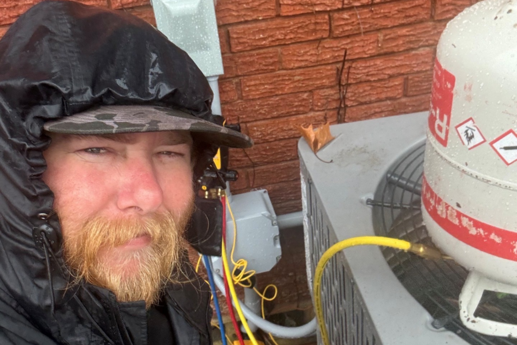 A close-up selfie of an HVAC technician with a blonde beard wearing a black rain jacket and a camouflage hat. In the background, he is servicing an outdoor AC unit with refrigerant gauges and a tank during a rainstorm.