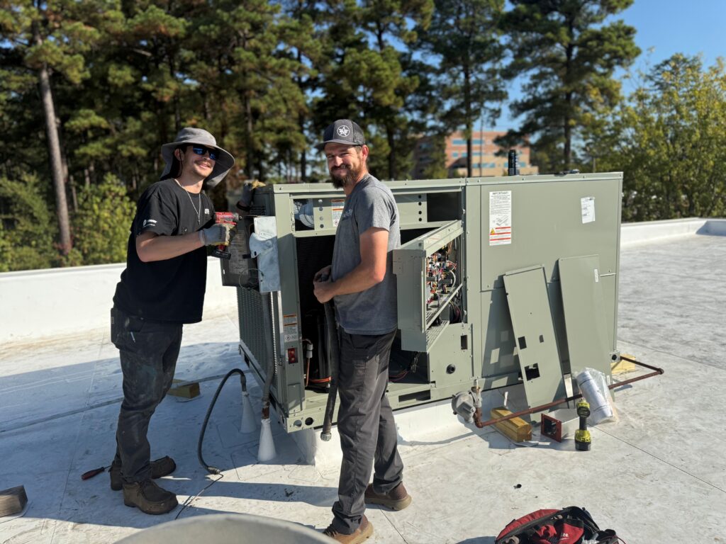 Two HVAC technicians smiling while installing a large rooftop unit on a flat white roof.